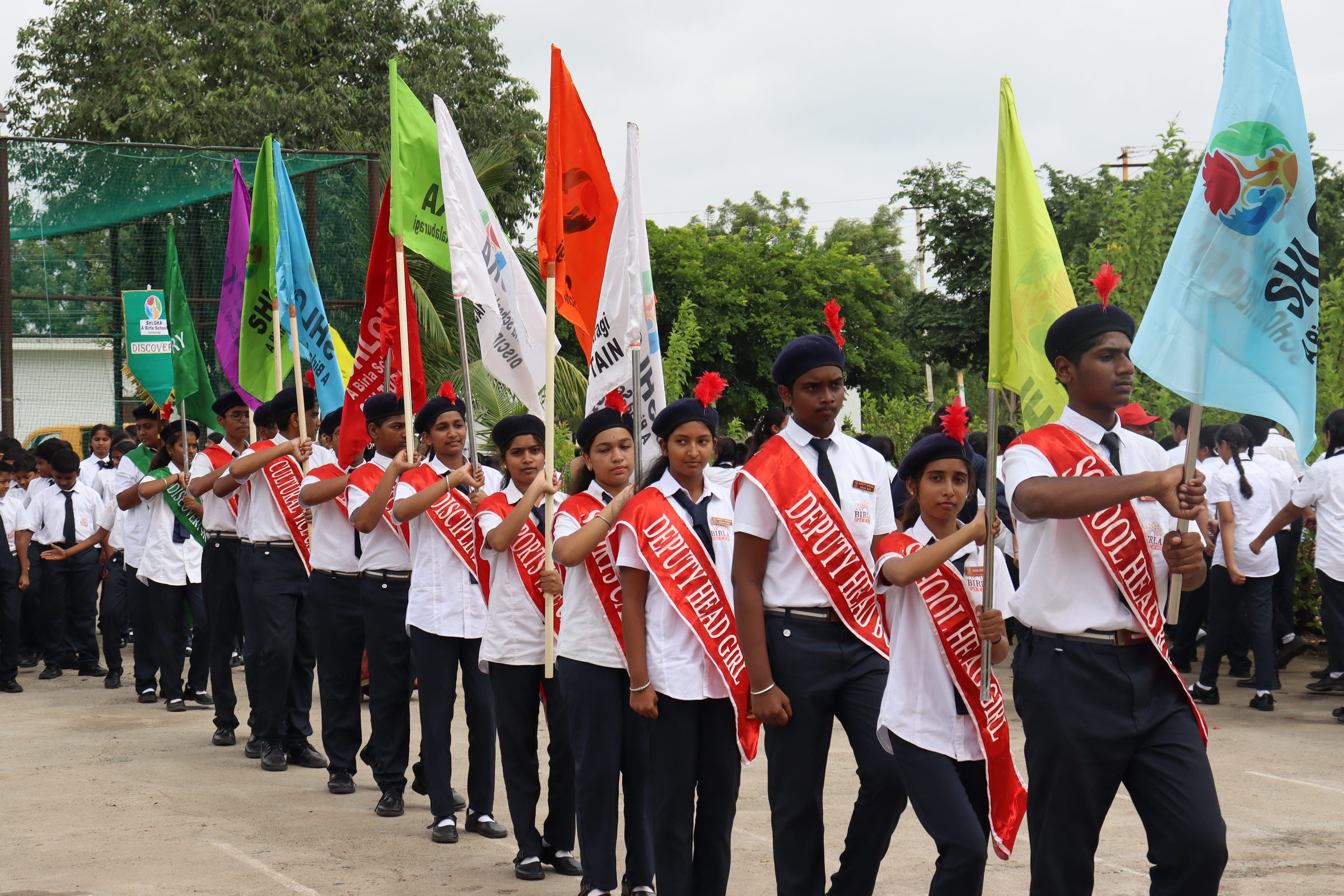 Students in school uniform and office sashes carrying house flags in the march past on the school grounds
